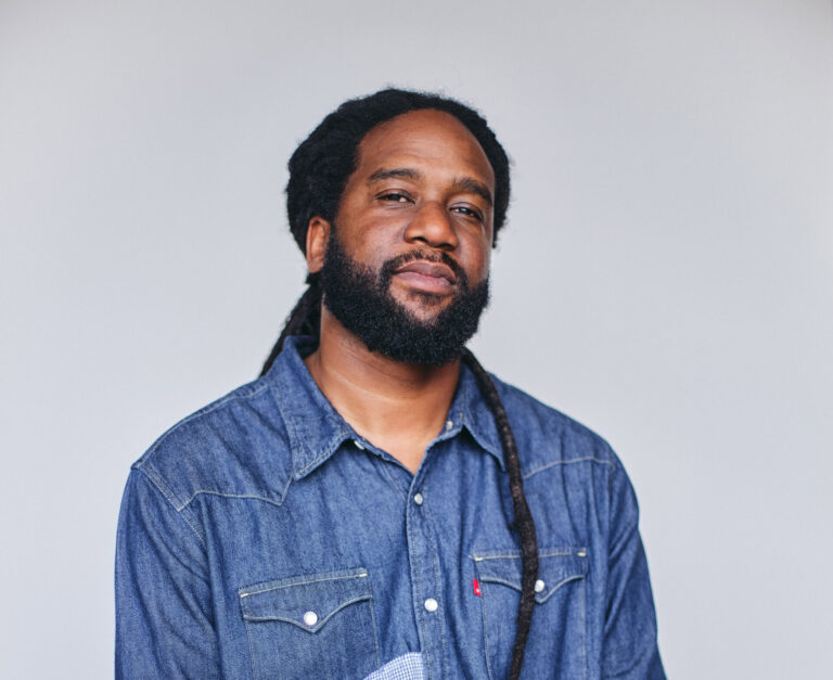 Donkere man kijkt in camera, gekleed in stoer spijkershirt, mouwen beetje opgestroopt waardoor tattoos op beide armen zichtbaar zijn