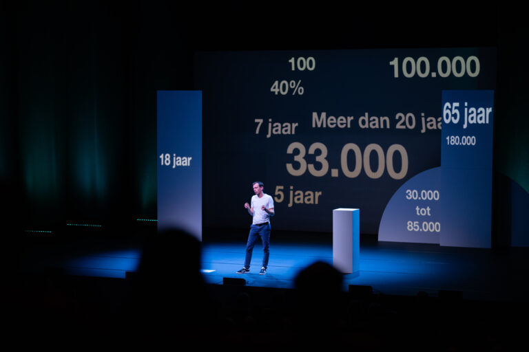 Een spreker staat op het podium voor een grote presentatie met verschillende cijfers en teksten op een scherm.