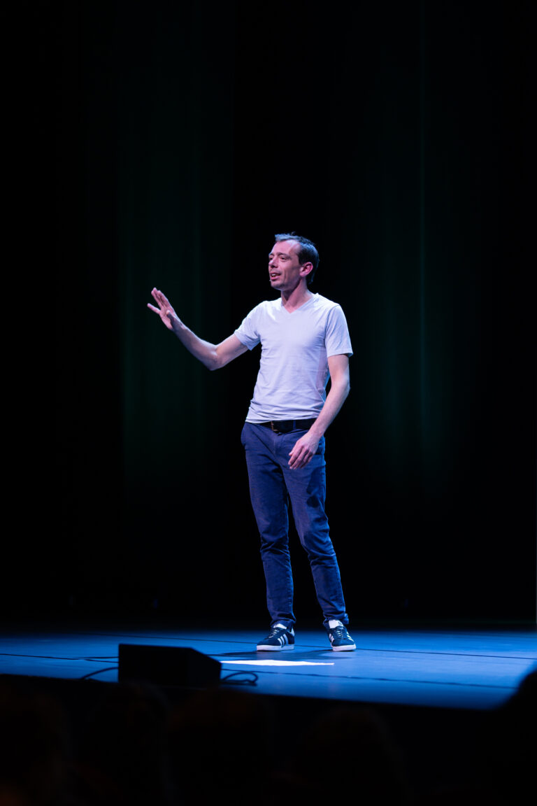 Een man in een wit T‑shirt en blauwe jeans staat op een donker podium en maakt een gebaar met zijn hand.