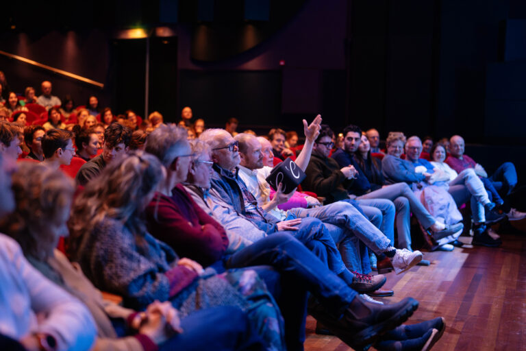 Een publiek zit op banken in een theater met een sfeervolle belichting, overweldigend door de donkere achtergrond.
