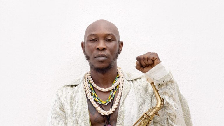 Seun Kuti poseert met saxofoon en heft zijn vuist in een krachtige, iconische pose.