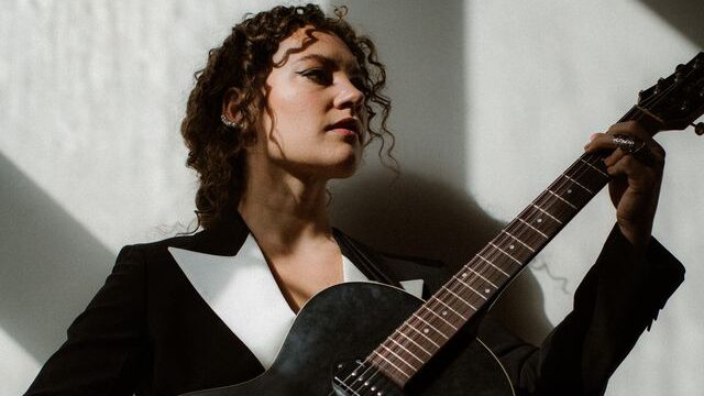 Persoon met gitaar in een zwart-wit outfit, kijkt omhoog naar het licht.