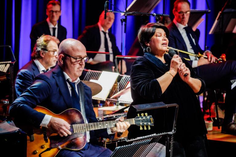 Een zangeres en een gitarist treden op met een orkest op de achtergrond.