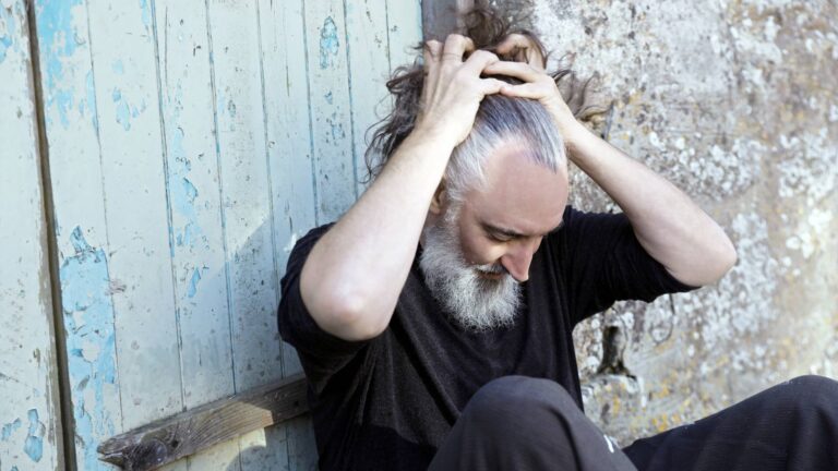 Bebaarde man met lang haar, zittend en zijn haar vastzettend tegen een verweerde muur