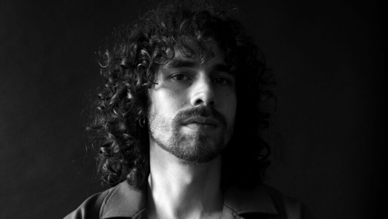 Zwart-wit portret van een man met krullend haar en een baard, kijkend in de camera.