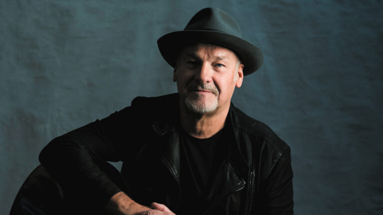 Paul Carrack, Britse zanger, met hoed en zwarte jas, tegen een donkere achtergrond.