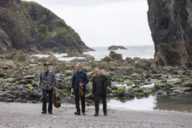 De drie mannen van Miscellany of Folk staan voor de zee. Er is een rotspartij te zien in de achtergrond. Een man heeft een gitaar, een ander een accordeon en de laatste heeft een viool. Ze kijken naar de grond of naar elkaar.