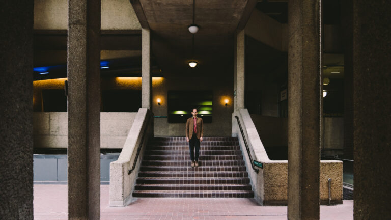 Een man staat onder een overdekte betonnen structuur bovenaan een brede trap, omringd door pilaren en warme verlichting.