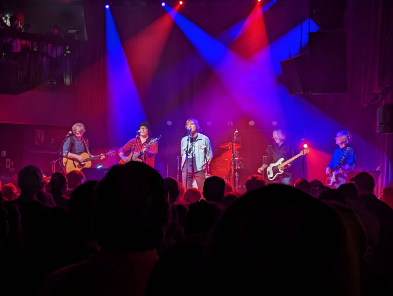 Band staat op het podium met rode en blauwe lichten achter zich. Publiek kijk je op de rus.