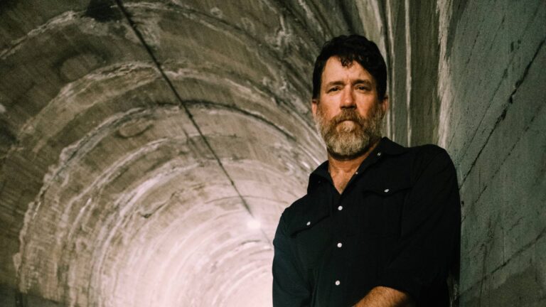 De afbeelding toont een man met een baard die een donkere, casual blouse draagt. Hij staat in een tunnel met betonachtige muren, die licht gebogen zijn. De verlichting van de tunnel creëert een dramatisch effect met schaduwen en contrasten op de achtergrond. De man kijkt serieus in de camera, met een rustige maar vastberaden houding.