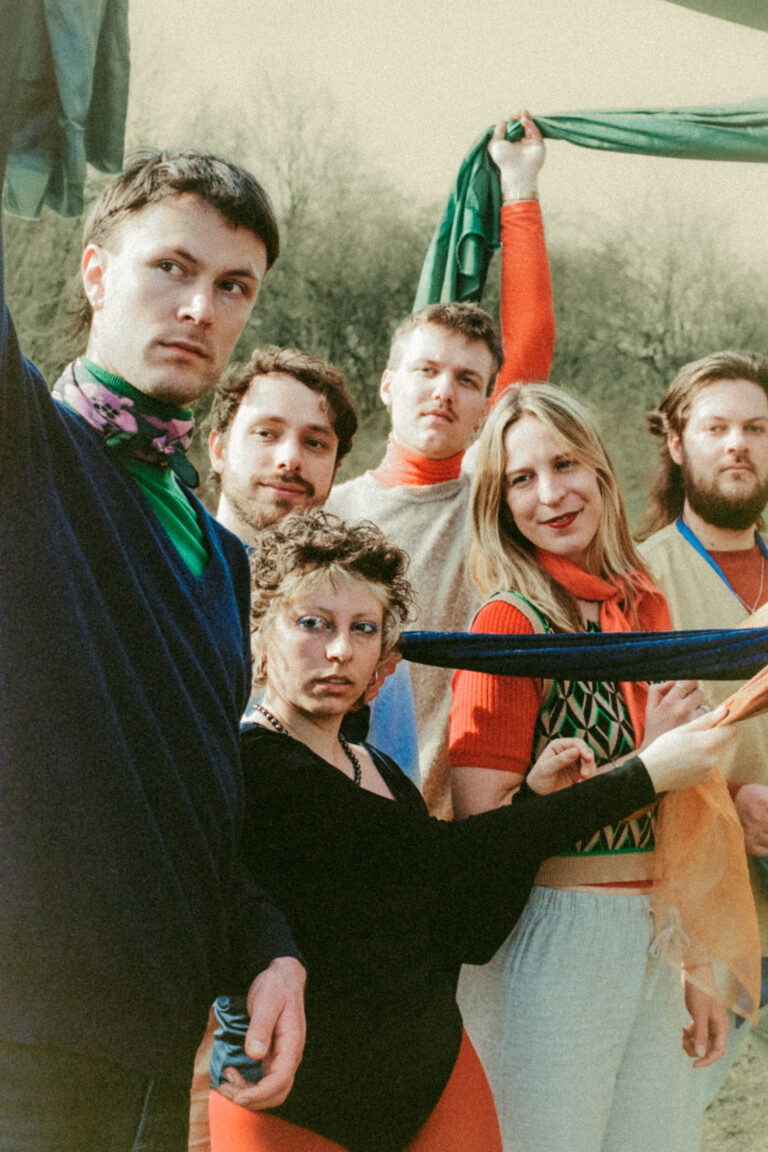 Een groep van zes mensen poseert buiten met kleurrijke kleding en accessoires.