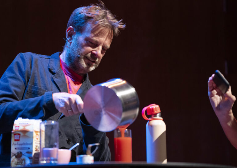 Teun van de Keulen schenkt een rode sap van een pan over naar een glas. Iemand maakt een foto met een telefoon van hoe hij dit doet.
