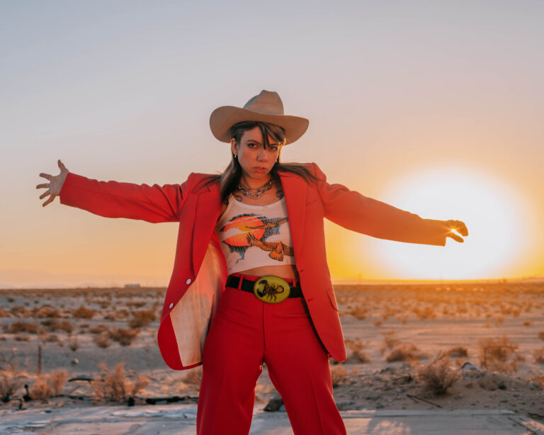 Vrouw met rood pak en cowboyhoed en armen wijd kijkt de camera in, achter haar ondergaande zon