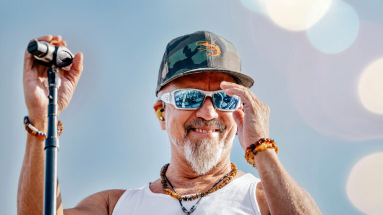 Een man met een grijze baard en zonnebril, gekleed in een wit hemd en camouflagepet, glimlacht terwijl hij op een podium staat en een microfoon vasthoudt