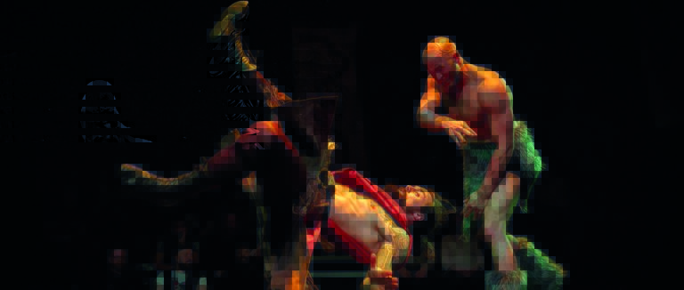 Een gespierde man tilt een andere man in een rode mantel op tijdens een theaterperformance. Het publiek kijkt toe op de achtergrond.