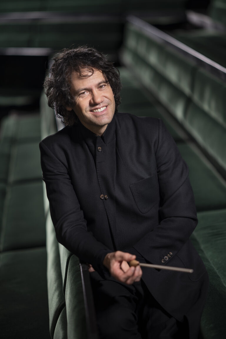 Een man in een zwart pak zit op een groene bank met een dirigeerstok in zijn hand.