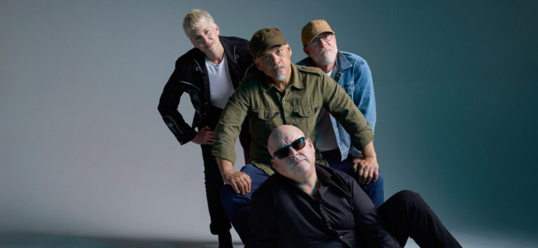 Vier personen poseren, één zittend op de grond, de anderen leunen voorover in casual kleding.