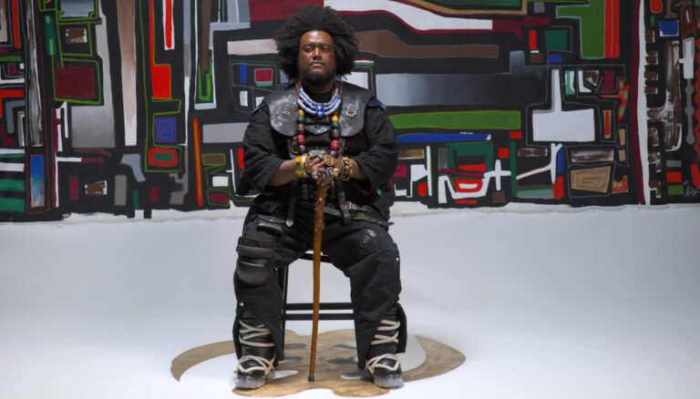 Een man zit op een stoel voor een kleurrijk abstract schilderij, met een wandelstok in zijn handen.