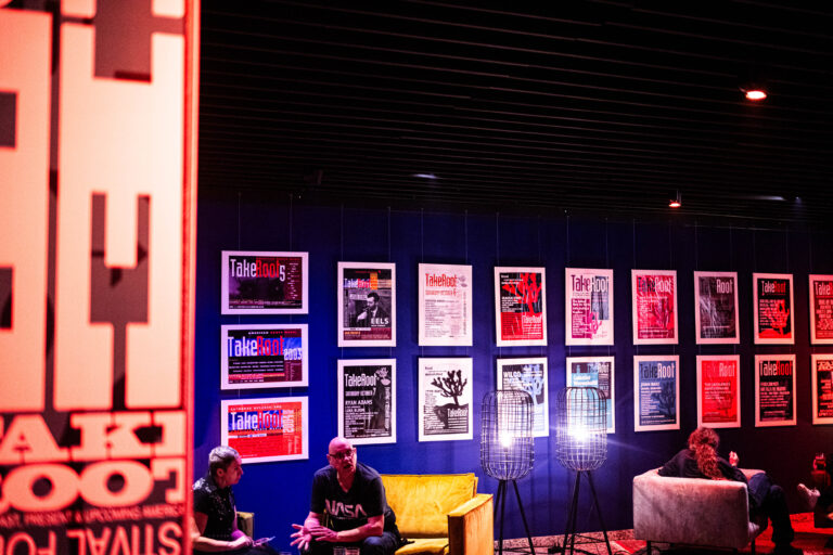 Wand met 25 line-up posters van TakeRoot op blauwe muur, ervoor stoelen met mensen erin