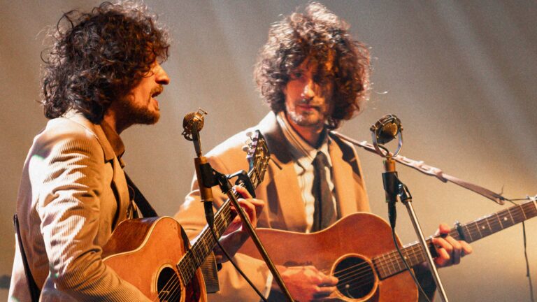 Twee muzikanten met krullend haar spelen gitaar op het podium met microfoons voor zich.