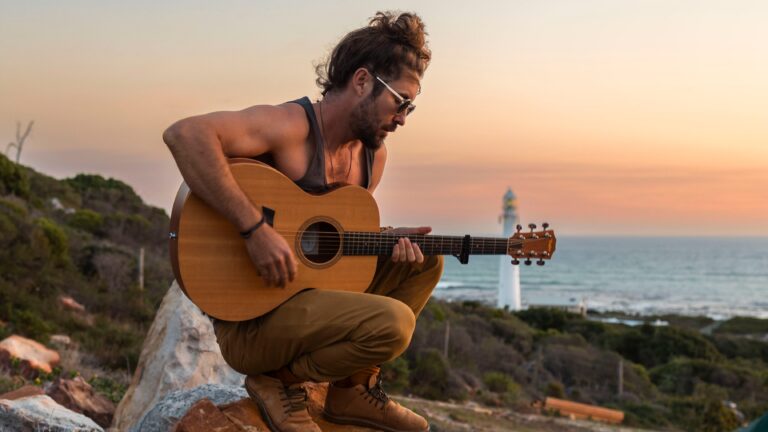 Man met een gitaar speelt muziek bij zonsondergang met een vuurtoren op de achtergrond.