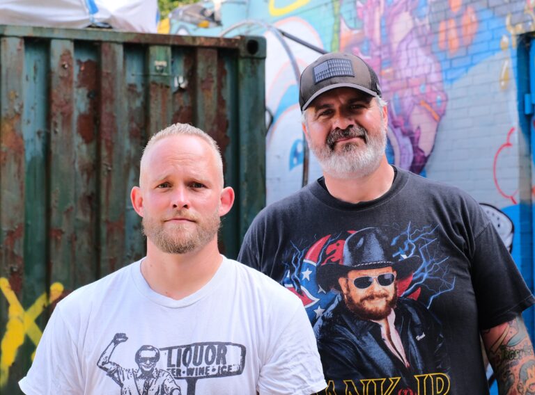 Twee mannen (Peter Kwint en Peter Bootsman) voor een kleurrijke graffiti achtergrond. Ze staren glimlachend de camera in.