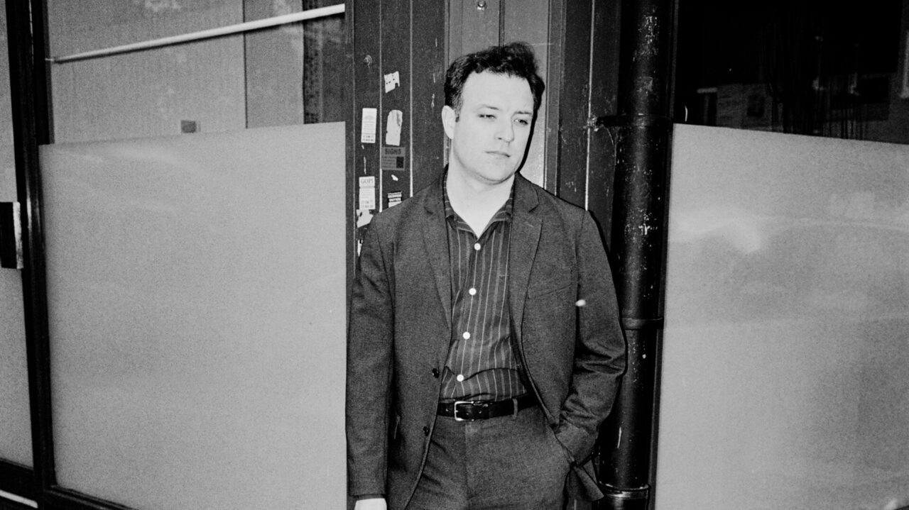 Zwart-wit portret van PM Warson, casual gekleed in een pak en leunend tegen de hoek van een gebouw op een stedelijke locatie.