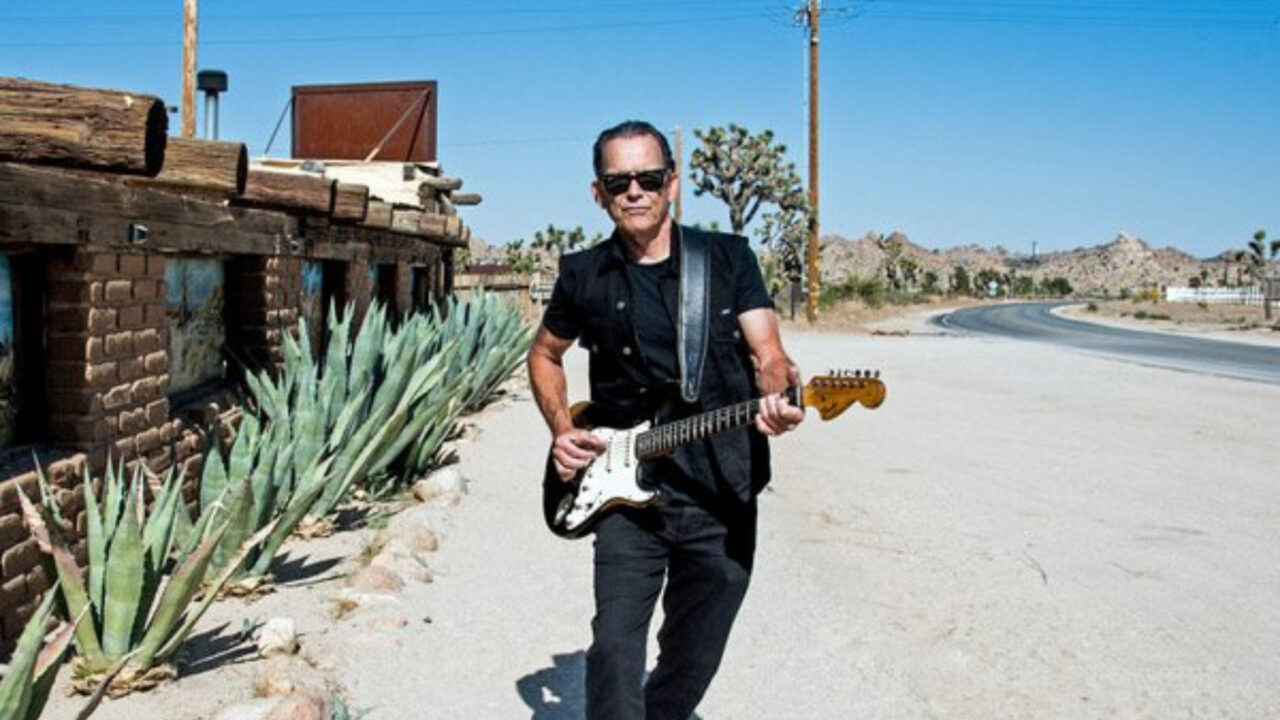 Tommy Castro staat buiten in een woestijnlandschap naast een stenen gebouw, terwijl hij een elektrische gitaar bespeelt en een zonnebril draagt.