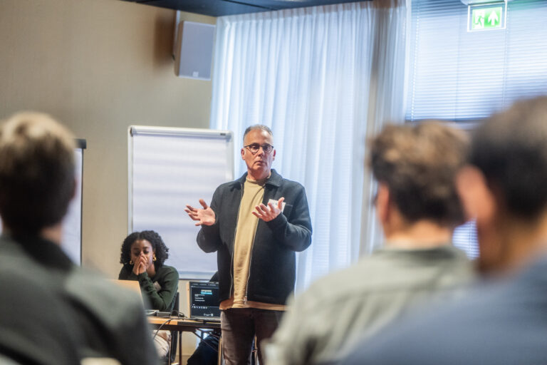 Een man met een bril geeft een presentatie in een zaal en maakt een gebaar met zijn handen. Achter hem zijn een flip-over en gordijnen zichtbaar. Een vrouw zit achter een tafel met een laptop en luistert aandachtig.