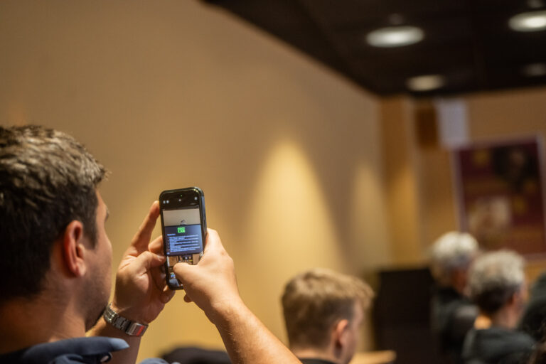Een man maakt een foto met zijn smartphone tijdens een presentatie. Het scherm van de telefoon toont een dia met tekst en een groen element. Op de achtergrond zitten andere mensen in een zaal met zachte verlichting.
