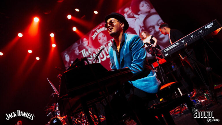 Een muzikant in een blauwe jas speelt enthousiast op een toetsenbord onder kleurrijke verlichting tijdens een optreden.