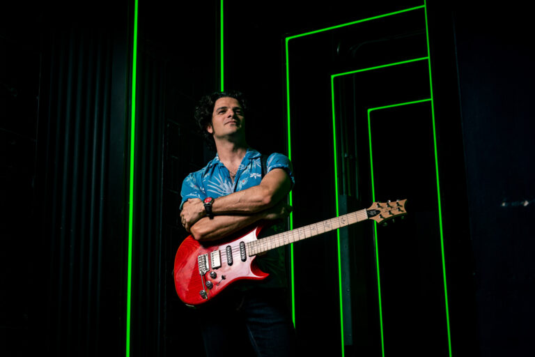 Mark Lettieri staat met zijn gitaar om zijn nek en zijn armen over elkaar. Hij staat in een soort zwarte kubus met groen neon licht.