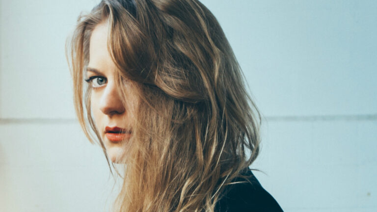 Jo Harman kijkt met een intense blik en losse, golvende haren over haar schouder in een lichte, neutrale achtergrond.