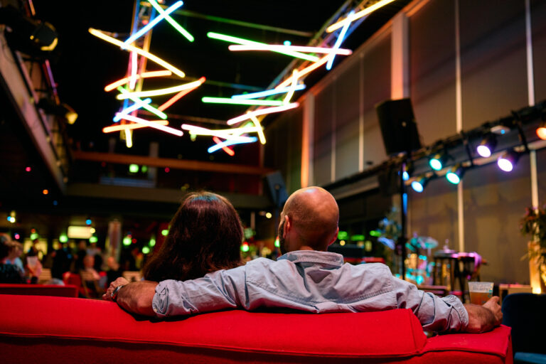 Man en vrouw die op een rode bank zitten te luisteren naar een band terwijl de man een arm om de vrouw haar schouders heeft geslagen.