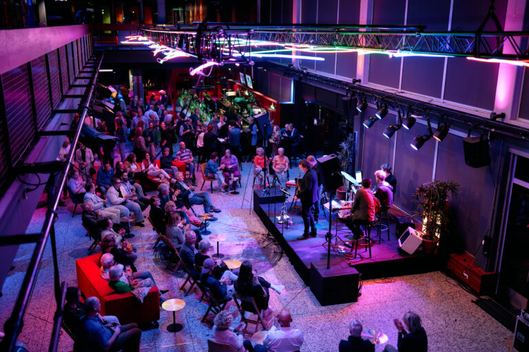 foto van bovenaf waarin we kijken naar the jazzmob die staat te spelen in de foyer. Met allemaal mensen die er omheen zitten te luisteren.