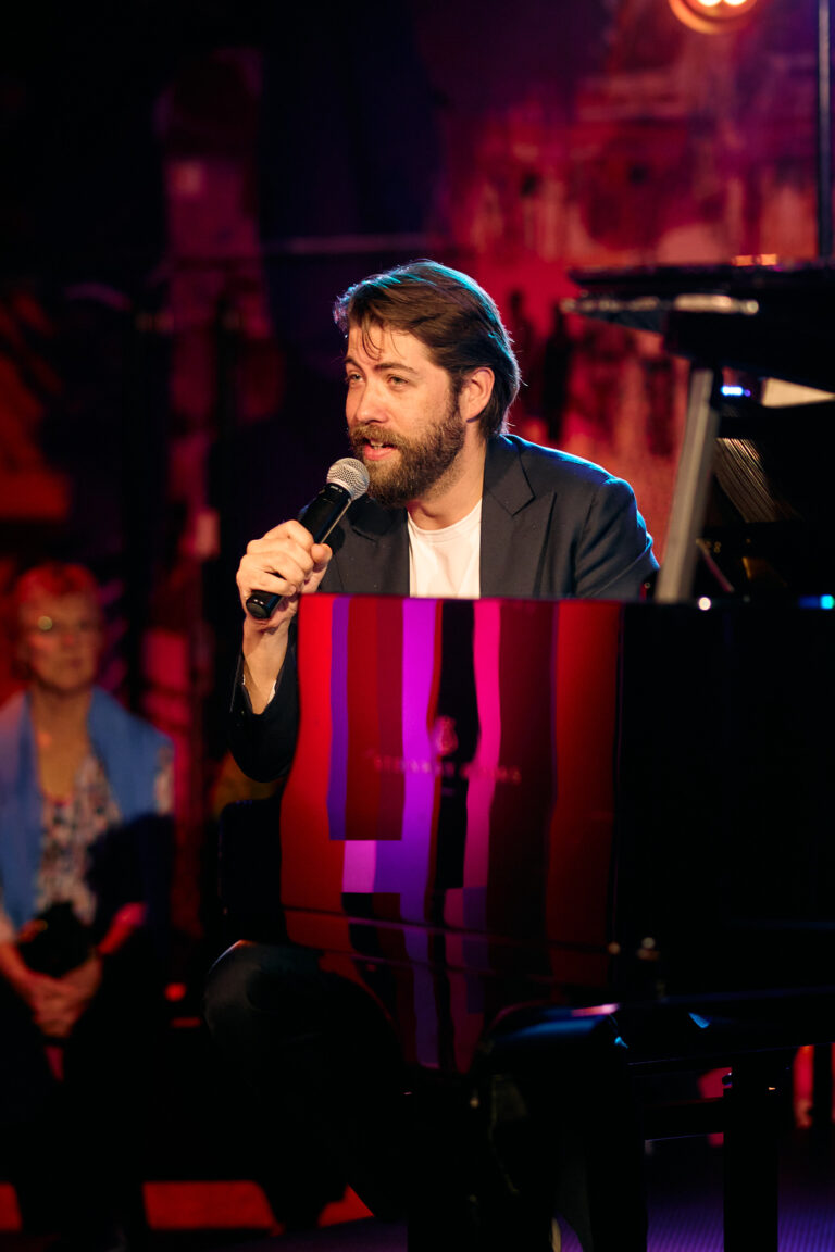 man met een microfoon in zijn hand die achter een vleugel zit en aan het vertellen is tegen een groep mensen