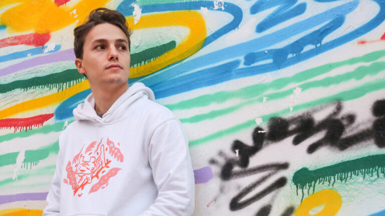 Matteo Mancuso staat nonchalant tegen een muur met veel kleuren erop. Hij heeft een witte trui aan en zijn handen in zijn zakken.