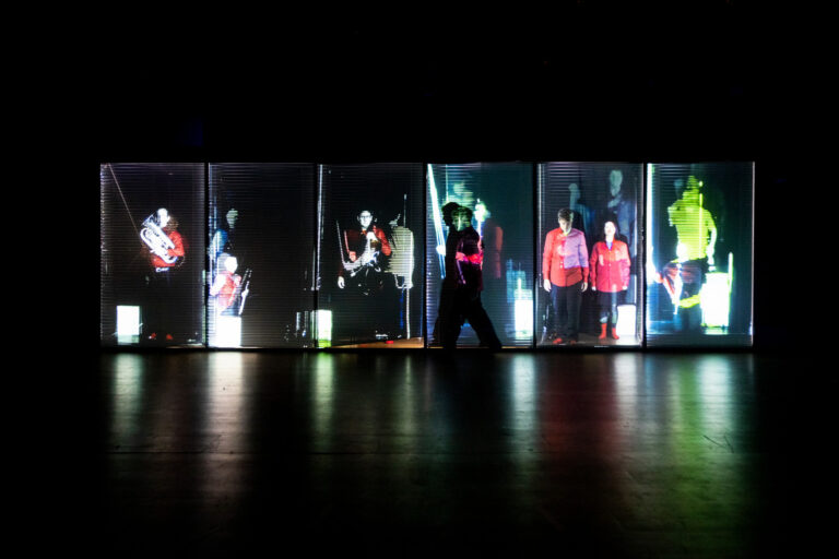 Muzikanten van het Nadar Ensemble, verspreid over zes met half open afgesloten lamellen. Op de lamellen zijn ook videoprojecties te zien, wardoor niet duidelijk is wie de echte muzikant is wie video is.