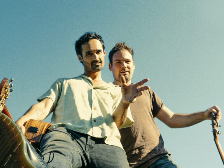 Twee muzikanten poseren met instrumenten onder een heldere blauwe lucht.