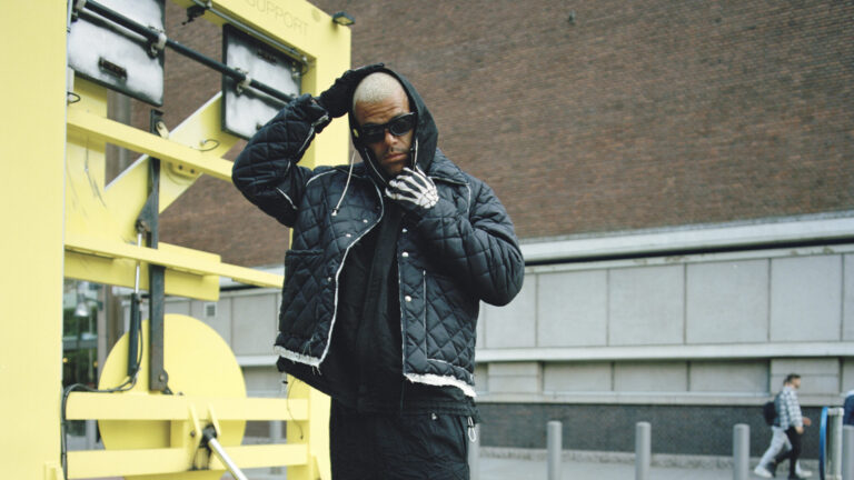 Een man in een zwarte quilted jas en zonnebril staat buiten naast een grote gele constructie.