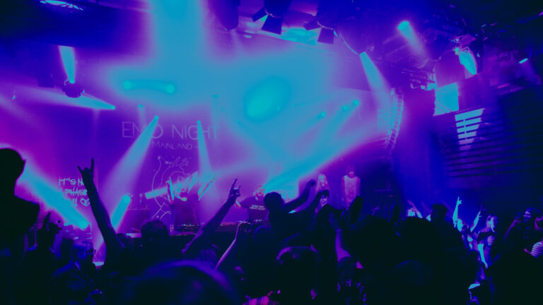 Een drukke concertzaal verlicht door paarse en blauwe lichten, met een enthousiaste menigte en artiesten op het podium tijdens een optreden.