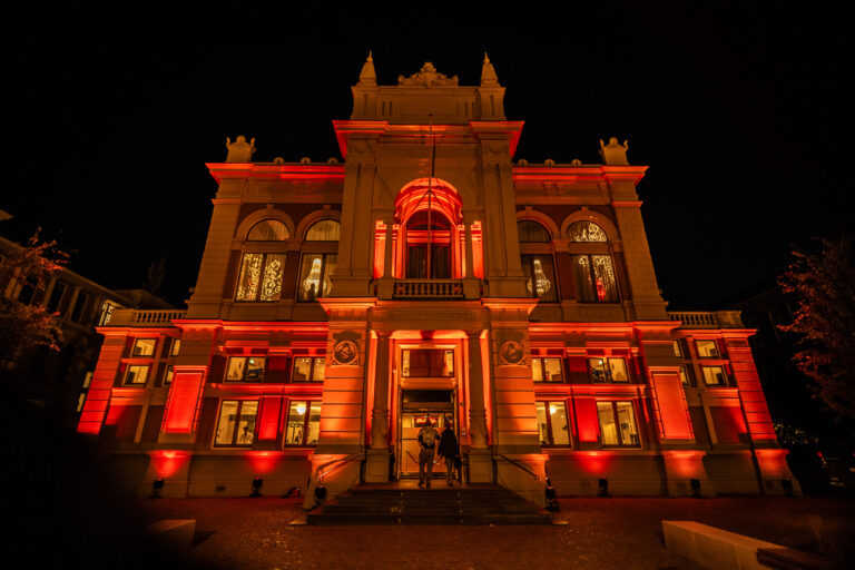Een indrukwekkend gebouw, verlicht met warme rode en oranje tinten, staat prominent in de nacht. Grote ramen tonen binnenin kroonluchters en decoratieve details. Twee mensen staan bij de ingang.