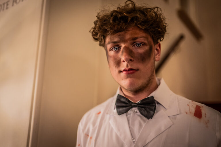 Een jonge man met krullend haar en intense blauwe ogen draagt een wit laboratoriumjas met bloedvlekken en een zwarte vlinderdas. Zijn gezicht is bedekt met roetvegen, wat een dramatische en mysterieuze uitstraling geeft.