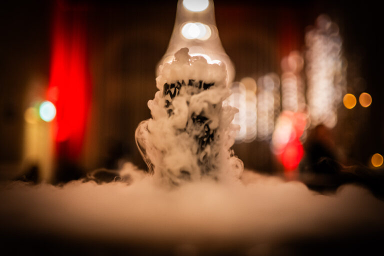 Een decoratief object omhuld door dichte witte rook staat centraal in een sfeervolle setting. De achtergrond toont onscherpe warme lichten en rode accenten, wat een mysterieuze en feestelijke sfeer creëert.