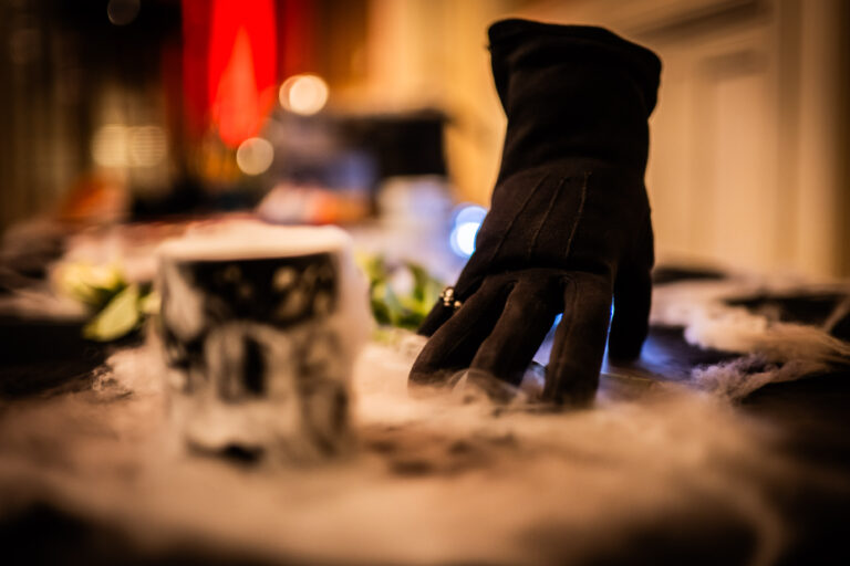 Een zwarte handschoen ligt op een tafel bedekt met decoratief spinrag. Op de voorgrond staat een zwart-witte mok, terwijl de achtergrond onscherpe warme verlichting en rode accenten toont, wat een mysterieuze en sfeervolle uitstraling creëert.