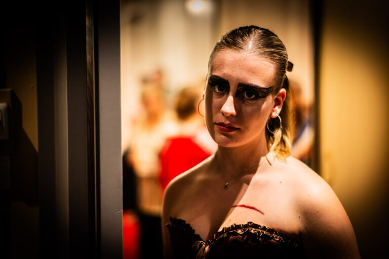 Een jonge vrouw met dramatische zwarte oogmake-up en strak naar achteren gekamd haar kijkt recht in de camera. De achtergrond is onscherp, met vage silhouetten van mensen in een verlichte ruimte.