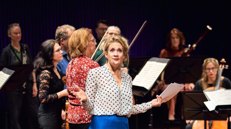 Jet Berkhout in blauwe broek en blouse voor orkest kijkt vragend naar publiek in de zaal met armen open.