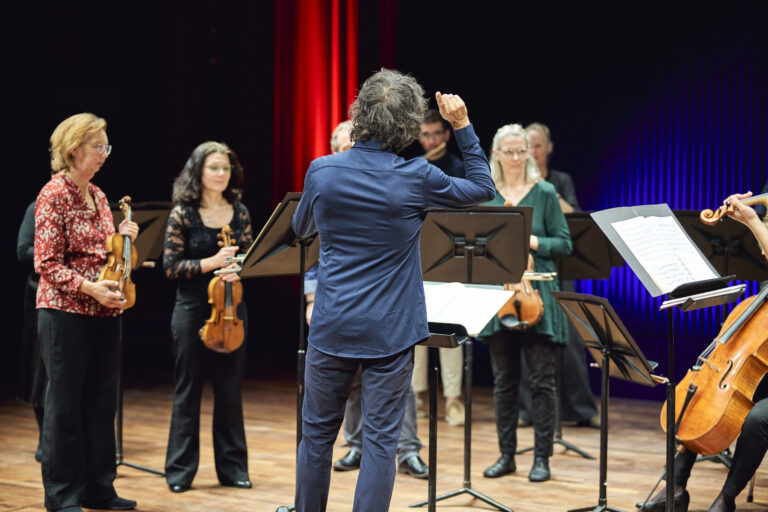 Dirigent die een grop mussici aan het dirigeren is. Met op de achtergrond violisten