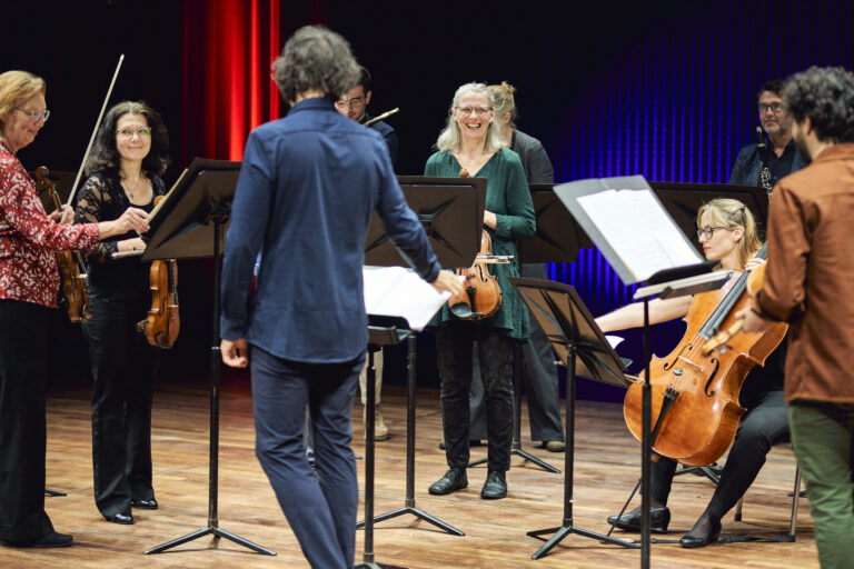 Dirigent die een grop mussici aan het dirigeren is. Met op de achtergrond violisten