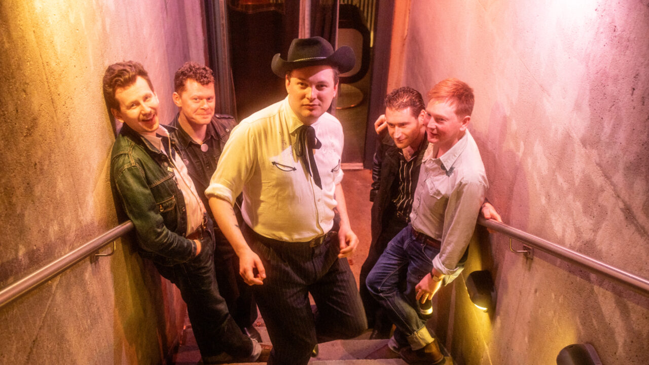 Rob Heron & The Tea Pad Orchestra in rockabilly-kleding, met één man in het midden die een cowboyhoed draagt, poseert op een trap in een sfeervolle ruimte met zachte, gekleurde verlichting.